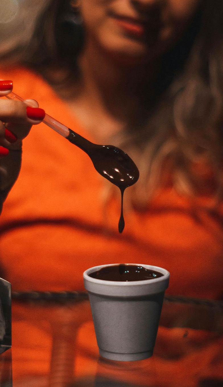 Una mujer en segundo plano sostiene una cuchara de la que cae lentamente chocolate caliente y espeso, justo sobre una taza blanca repleta del mismo. El primer plano se centra en la textura sedosa del chocolate y en la calidez del momento, mientras el fondo desenfocado y el tono naranja de su ropa aportan una sensación de confort, placer y disfrute.